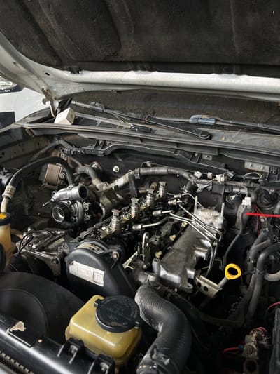 The image displays the engine bay of a vehicle, in a state of disassembly for maintenance, located in La Peña, Panama. The vehicle's hood is open, revealing a multi-cylinder diesel engine. A turbocharger unit is detached and rests on the engine block, left of the cylinder head. Four fuel injectors and a common rail are visible on the cylinder head, with signs of oil and grime accumulation. A black timing cover and a visible serpentine belt are present. Various hoses, wires, and electrical connectors are routed throughout the compartment. A translucent yellow power steering fluid reservoir with a black cap is in the lower left foreground. An oil dipstick with a yellow loop is situated on the right side of the engine. A small, light-colored cardboard box rests on the dusty cowl panel, which features two windshield wiper blades. The underside of the hood is covered with black insulation. A red arrow, an external annotation, points to a white electrical connector on a wiring harness on the right side of the engine bay.