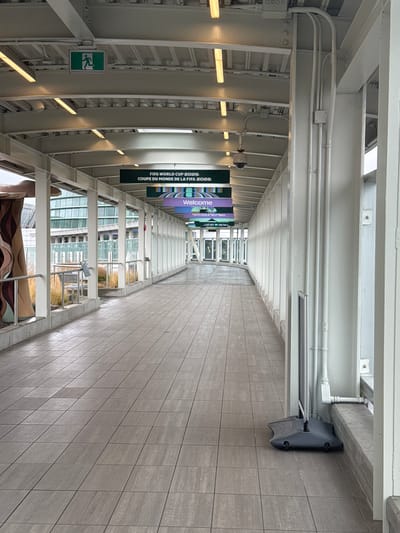 This is a vertical, eye-level, medium shot of an outdoor walkway or concourse, likely in Richmond, Canada. The walkway is covered by a white, slatted roof, supported by a metallic structure. The floor is tiled with large, grey, rectangular tiles. The right side of the walkway is lined with a tiled wall.

Banners hang from the overhead structure, with the visible text "FIFA WORLD CUP 2026 COUPE DU MONDE DE LA FIFA 2026" and "Welcome." The banners suggest the event or a related celebration is underway or upcoming.

On the left side of the walkway, an illuminated green exit sign with a running figure is visible. Further down the walkway, several tables and chairs are arranged, suggesting a seating or waiting area. The lighting appears to be artificial, with fluorescent fixtures casting a warm glow. The scene is bright, indicating it could be daytime.

There are no people visible in the immediate foreground or midground. The overall impression is one of modern public infrastructure, possibly at an airport, train station, or convention center, decorated for a major sporting event.
