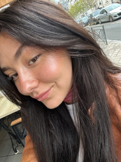 The image shows a close-up of a woman's face. The woman is looking down and to the right. She has long, dark hair and is wearing a brown leather jacket. She appears to be smiling slightly.  The background of the image includes the lower part of the woman's white shirt and a blurry view of a city street.  The woman's nose is the primary focus of the photo.  The coordinates provided (latitude: 47.587, longitude: 1.337) indicate a location in Blois, France, but the image does not offer any visual clues to confirm this.