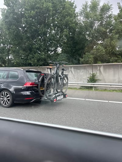 A black Volkswagen Golf station wagon is parked on the right side of a paved road, likely a highway, with two bicycles attached to a rear-mounted bike rack. The car is viewed from the rear right. Trees line the background beyond a concrete sound barrier. The sky is overcast. The license plate of the car is visible and reads "ZT-599-R". One of the bicycles is a dark color with silver accents and appears to be an electric bike. The other bicycle is partially obscured. The scene is typical of a highway in or near Harderwijk, Netherlands, given the Dutch license plate and the common practice of transporting bicycles for recreation in such areas. There are no people visible in the immediate vicinity. The road surface is asphalt with white lane markings.