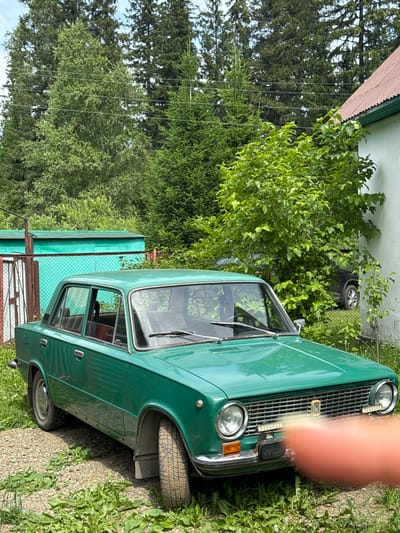 The image depicts a green, vintage sedan parked on a gravel and grass surface in what appears to be a rural or suburban setting. The car is the primary subject, positioned centrally and facing slightly to the left. In the background, there are tall evergreen trees, a portion of a white building with a reddish-brown roof, and a teal-colored metal shed with a red-brown fence in front of it. A dark-colored car is partially visible behind a bush to the right of the sedan. The sky is visible through the trees, suggesting it is daytime. The overall scene is one of a quiet, outdoor environment.