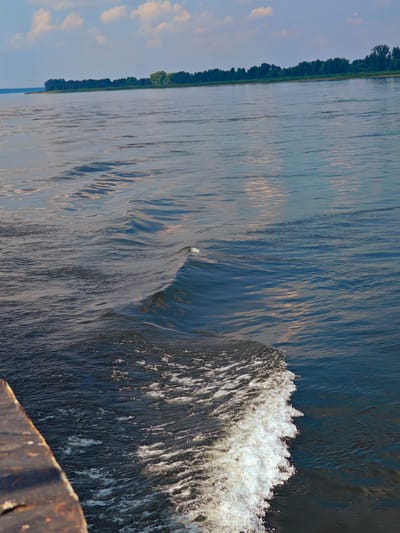 The image shows a wide river with ripples and a small wave breaking on the right side. The water is dark blue and reflects the sky, which is partly cloudy and blue. In the background, a tree line runs along the distant shore, suggesting a natural landscape. The foreground includes a dark, weathered wooden surface, possibly part of a boat or dock, on the lower left. The scene appears to be a peaceful, natural setting. The location of Ust'-Kem', Russia, is not depicted or suggested in the image.