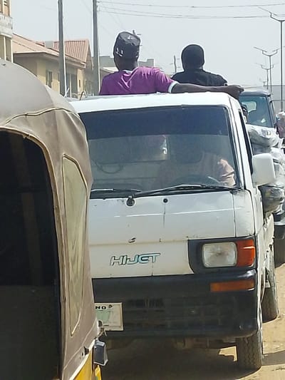 The image depicts a daytime street scene in Fagge D1, Nigeria. In the foreground, the tan canvas canopy and side of a three-wheeled vehicle are partially visible on the left. Directly behind it, a white Daihatsu Hijet pickup truck is positioned centrally. The truck features a black front bumper, a visible right headlight, and a partially obscured Nigerian license plate reading "NO" on its lower front fascia. A driver, wearing a light-colored garment, is faintly visible through the windshield. Two individuals are riding upright in the bed of the Hijet, positioned behind the cab and visible from the waist up. The person on the left wears a purple short-sleeved shirt with white trim and a dark cap, looking towards the right. The person on the right wears a dark short-sleeved shirt and has their left arm resting on the left individual's shoulder; they are also looking right. In the background, another vehicle carrying bundles or sacks is visible behind the Hijet. Buildings with light-colored walls and reddish-brown sloped roofs line the left side of the street. Utility poles and overhead power lines are present, along with streetlights on the right. The ground appears unpaved and dusty under a clear, bright sky.