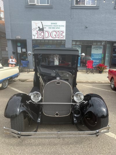 A vintage black Ford Model A coupe is parked on a paved street in front of a building with businesses named "The EDGE" dance studio and "CLEAN MY HOME" cleaning services. The building is grey and has the address 4816B visible above a closed door. A woman sits in a chair outside the studio, looking at her phone. Red chairs and potted flowers are visible outside the cleaning service business. The car has a chrome grille, round headlights, and a visible horn. The background shows more businesses and a glimpse of other vehicles. The scene appears to be in the City of Leduc, Alberta, Canada, based on the phone number 780-986-5252 displayed on "The EDGE" sign, which is a local area code for Leduc.