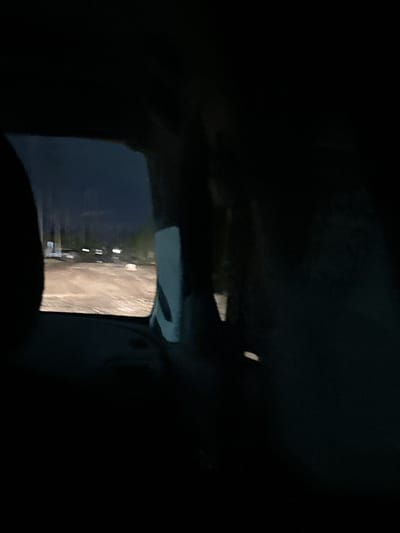 The image depicts a nighttime scene, viewed from inside a vehicle, looking out the rear window. The exterior environment appears to be a rural or semi-rural area, possibly a road or open lot illuminated by artificial light sources. In the distance, faint rectangular shapes suggest buildings or structures with windows emitting light. The foreground is dominated by the dark interior of the vehicle, including the dark shapes of seats or interior panels. The road surface is visible as a lighter, textured area, reflecting the ambient light. The overall impression is of movement or stillness at night in a sparsely populated area. Without further information or visual cues, it is impossible to definitively place the scene within Заречный ауылдық округі, Kazakhstan.