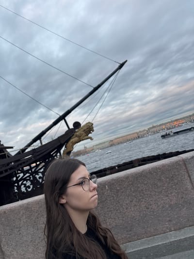 A young woman with long brown hair and glasses stands on a stone embankment, looking to her right. Behind her, a portion of a ship's dark hull is visible, adorned with a golden lion figurehead. The ship is docked in the Neva River in Saint Petersburg, Russia. In the background, across the river, a row of historic buildings stretches along the embankment, illuminated by lights. The sky is overcast with dramatic clouds, and several small boats are visible on the water.
