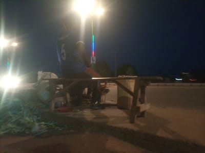 A person wearing a blue shirt with the number 5 on the back is seated on a motorcycle at night in Wuro Dole, Nigeria. The person is illuminated by a bright street light. In the foreground, there is a wooden table with some items on it, and to the left of the table, there is a pile of green material, possibly produce or fishing nets. The background is dark with a hint of the night sky. The overall scene appears to be outdoors, possibly near a market or roadside stall.