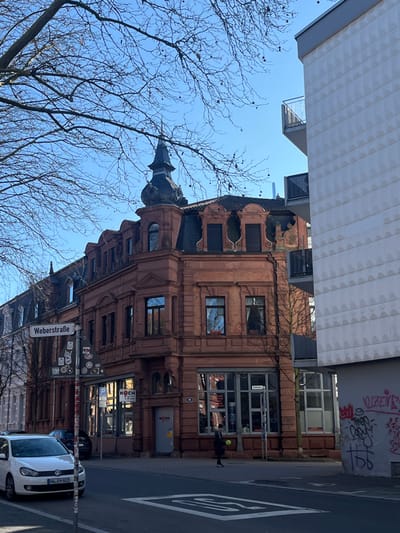 The image depicts an urban street scene at Weberstraße in Kaiserslautern, Germany, under a clear blue sky. Bare tree branches are visible in the upper portion of the frame.
The central feature is a multi-story building on a corner, constructed with a reddish-brown brick facade and a dark gray mansard roof. It features an ornate corner tower with a spire. The ground floor includes large display windows and an entrance with a "KOCH" sign. Adjacent to this building on the right is a modern, multi-story structure with a light-colored, textured facade and dark gray balconies with metal railings. The lower section of this modern building's wall exhibits graffiti. An additional older building is partially visible to the left of the historic corner building.
A white Volkswagen Golf (license plate KL-EM 602) is parked on the left side of the street. White road markings, including an arrow and a crosshatch pattern, are visible on the asphalt. A street sign pole in the left foreground displays "Weberstraße" and various stickers.
A single individual, dressed in dark clothing, is observed walking on the sidewalk in front of the historic building. This person is holding a bright yellow, spherical object in their left hand.