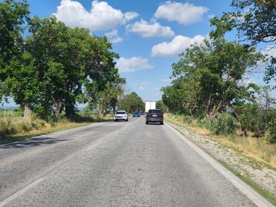 The image depicts a road in Totleben, Bulgaria, on a clear day with scattered clouds in the blue sky. The road is paved and appears to be a two-lane highway, with a white line marking the center. Trees line both sides of the road, creating a canopy overhead in some sections. Several vehicles are present: a white Mercedes-Benz sedan, a blue sedan, a black SUV, and a white truck further down the road. The vegetation along the roadside consists of grass and bushes. The overall scene suggests a journey through a rural or semi-rural area.