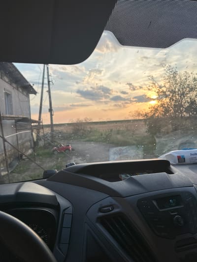 The image is taken from inside a vehicle, looking out the windshield towards a rural landscape at sunset. The sky is filled with dramatic clouds illuminated by the setting sun, casting a warm, orange glow over the scene. In the foreground, the dashboard of the vehicle is visible, including a steering wheel, an instrument cluster with a speedometer, and a car stereo system.
Outside the vehicle, to the left, is a simple, light-colored building with a sloping roof. Adjacent to it is a utility pole. Further into the scene, on the ground, is a small red toy car. The landscape consists of fields and sparse vegetation, with a few trees visible in the distance. The windshield has several specks of dust or dirt on it, which are illuminated by the sunlight.
The presence of the toy car and the rural setting suggest this image might have been taken in Mlekarevo, Bulgaria, a village known for its agricultural surroundings. However, without further information, this is an assumption based on the visual cues.
