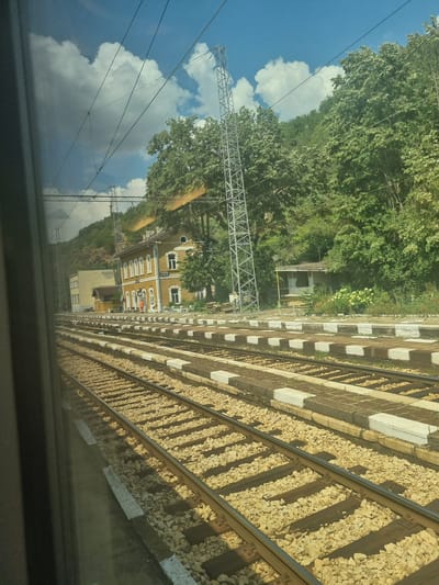 The image shows a view from a train window, looking out at a railway station in Zlidol, Bulgaria. The foreground is dominated by multiple sets of train tracks, with ballast visible between the wooden sleepers. Beyond the tracks, a low, yellow building with a red-tiled roof and white trim serves as the station building. It features several windows and a sign reading "ZLATOL". A single person in an orange uniform stands near the entrance of the building. Adjacent to the station is a smaller, older-looking wooden structure. Behind the buildings, a dense green forest covers a hillside. Overhead, numerous power lines crisscross the frame, supported by tall metal pylons. The sky is blue with scattered white clouds. The overall scene depicts a functional, somewhat rustic railway station set against a natural backdrop.