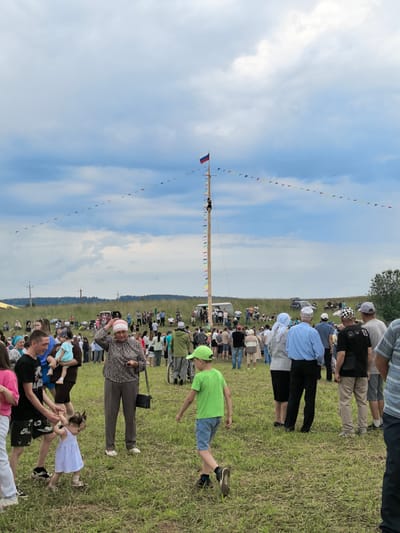 The image depicts an outdoor gathering of people in a grassy field, likely in or around the village of Koyanovo, Russia, given the presence of a Russian flag on a tall pole. A crowd of individuals of various ages is present, with some standing and others seated. A tall wooden pole is erected in the center of the scene, decorated with a string of colorful triangular flags. A person is visible climbing the pole. Several vehicles are parked in the background. The overall atmosphere suggests a festive or community event.