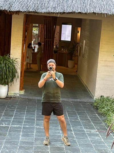 The image shows a man taking a mirror selfie in what appears to be a resort or hotel setting in Mu Cang Chai, Vietnam. The photograph is oriented vertically, with the man's body appearing horizontal across the frame.
The central subject is a man, wearing a green short-sleeved t-shirt, black shorts, a grey baseball cap, and light-colored sneakers. He is holding a black digital camera, pointing it towards the reflective surface to capture his own image. A watch is visible on his left wrist, along with a red bracelet.
The background reveals an inviting indoor-outdoor space. To the left, a textured thatched roof creates a natural, rustic architectural element. A dark grey tiled pathway or floor with distinct white grout lines stretches across the lower portion of the image. Potted green plants are strategically placed, enhancing the natural ambiance; one large plant in a white pot is prominent in the foreground.
Beyond the man's reflection, the interior space features warm-toned walls and intricate bamboo structures, suggesting traditional Vietnamese design. In the deeper background, two blurry figures are subtly visible, possibly reflections of other people or actual individuals within the space. A rectangular screen or monitor mounted on a wall displays light-colored text, which is illegible due to distance and resolution, but suggests an information display or menu.
The lighting indicates it is daytime, casting a bright but diffused light over the scene. The overall atmosphere is calm and peaceful, characteristic of a serene holiday destination.