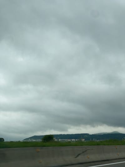 The image shows an overcast sky over Akhalsopeli, Georgia. A road with a white line marking the edge runs along the right side of the frame. A strip of green grass and trees runs along the road. Buildings are visible on the slope of a distant mountain.