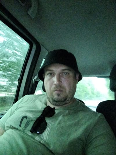 The image shows a man wearing a black bucket hat and a light green t-shirt. He is sitting in the passenger seat of a vehicle. Sunglasses are resting on his t-shirt. The interior of the car is dimly lit, with the ceiling and part of the back of the driver's seat visible. Through the car window on the left, a blurry, green, outdoor scene is visible, suggesting foliage. The light filtering through the window indicates it could be daytime, possibly overcast or rainy due to the muted lighting and the slightly washed-out appearance of the outdoor scene. The man's facial expression is neutral. The overall mood is casual.