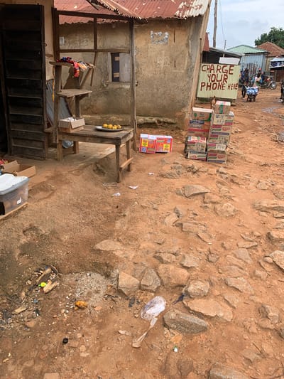 This image depicts an outdoor scene in Mape, Nigeria. In the foreground, there is a dirt ground covered with rocks and debris. To the left, a wooden table holds a silver plate with several yellow fruits. Next to the plate is a cardboard box. Behind the table is a wooden structure with shelves holding various items, including clothing and more cardboard boxes.

Further back, a weathered building stands with a corrugated metal roof. A sign on the building reads "CHARGE YOUR PHONE HERE" in red letters. A stack of cardboard boxes, appearing to contain food items, is placed near the building. In the background, several people and motorcycles are visible on a dirt road, suggesting a bustling marketplace or street. The overall impression is of a vibrant, albeit somewhat unkempt, urban environment.