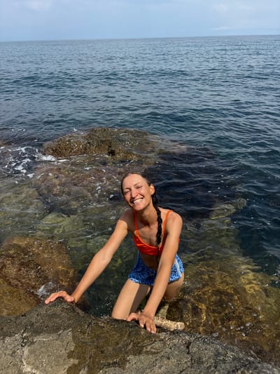 A woman is posing for a picture in the water near a rocky shore. She is wearing an orange bikini top and blue patterned shorts. She is smiling at the camera and holding onto a rock. The water is clear and the seabed is visible through the water. The sky is blue and there are some clouds. The location is the city of Utios, Ukraine.
