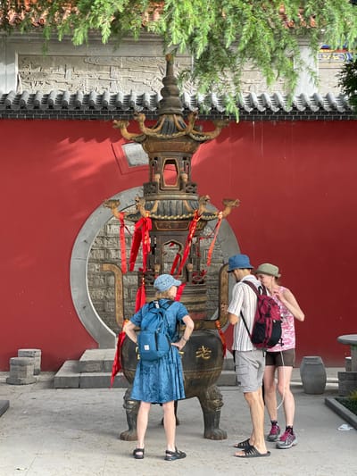In Lintong District, China, a photo captures a group of tourists near a large bell. The scene includes a woman in a blue dress and matching hat, and two other tourists inspecting the bell. The bell is adorned with red ribbons and intricate carvings. The backdrop consists of a red wall with a circular opening, behind which is a carved stone wall. A green tree stands to the left of the wall.