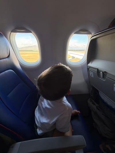 An overhead interior shot shows a child's head and shoulders as they look out of an airplane window. The child, wearing a white shirt, is seated in a blue airplane seat. The view outside the window reveals a sunny landscape with a mountain range in the distance, a tan-colored field, and the wing of the airplane. The airplane interior includes gray seatbacks with storage compartments visible. The image is not specific to any location in Donja Cijevna, Montenegro; it depicts a scene inside an airplane.
