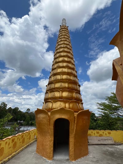 A golden, tiered stupa dominates the scene under a partly cloudy blue sky, located in Đăk Mar, Vietnam. The conical structure is adorned with multiple horizontal bands, each featuring a repetitive pattern of seated Buddha figures in relief. A white, sculpted figure crowns the apex of the stupa. At its base, a prominent golden structure with lotus-petal motifs forms an arched entrance into a darkened interior. The stupa stands on a flat, grey concrete platform. To the left, a low yellow wall or railing with stylized orange lotus designs extends along the platform's edge. On the far right, a curved golden architectural element of an adjacent building is partially visible. The background features lush green trees and distant vegetation, indicative of a natural, possibly elevated, setting. No individuals or specific actions are depicted.