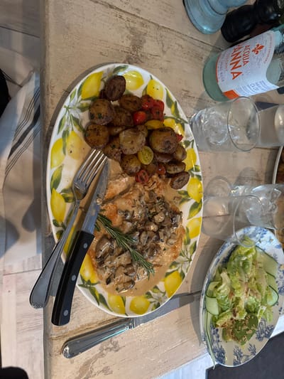 The image depicts an indoor dining scene, presumably located in Miami Beach, United States. A light-colored, possibly distressed wooden table serves as the primary surface. Centrally positioned is a large oval ceramic platter featuring a yellow lemon and white flower pattern. The platter holds two portions of light-colored meat, likely chicken, covered in a creamy mushroom sauce and garnished with a single sprig of fresh rosemary. Alongside the meat are roasted small potatoes mixed with red and yellow cherry tomatoes. A metal fork and a black-handled metal knife are placed on the left side of this platter. To the right, a smaller oval ceramic plate, adorned with a blue and white decorative pattern, contains a green salad comprised of lettuce, sliced cucumbers, and seeds, coated in a light dressing. In the upper right quadrant, an "Acqua Panna" natural spring water bottle is visible, accompanied by two clear glasses containing ice cubes. A striped fabric item, possibly a napkin or towel, in white and dark stripes, is partially visible on the far left. No people or specific actions are depicted.