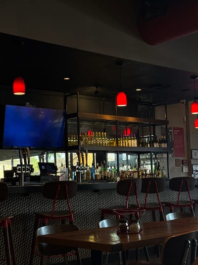 The image depicts the interior of a restaurant, specifically the bar and a portion of the dining area, identified as an Anthony's Coal Fired Pizza location in Aventura, United States. The scene is devoid of people.
The prominent feature is a well-stocked bar with a black counter. The bar's facade features a textured black and white geometric pattern. Several bar stools with dark wooden seats and red metal frames are positioned along the counter. Above the bar, a large flat-screen television displays a predominantly blue image. To the left, a digital point-of-sale system is visible on the counter. Two sets of beer taps are present: one to the left with four handles, including a "BUD LIGHT" tap, and another to the right with multiple handles.
Behind the bar, a multi-tiered metal-framed shelving unit holds numerous bottles of alcohol. The upper shelf contains rows of wine bottles, primarily white or light-colored. The lower shelf displays a variety of liquor bottles, including vodkas, rums, and whiskies, some illuminated from below. Hanging wine glasses are visible under a section of the shelving. A large window or mirror behind the bar reflects blurred outdoor light and shapes, suggesting foliage or buildings.
The ceiling is dark, with multiple recessed lights and three prominent red, bell-shaped pendant lights illuminating the bar and surrounding area. To the far right, a red rectangular sign titled "ANTHONY'S STORY" with an accompanying photograph is mounted on a light-colored wall.
In the foreground, a glossy dark wooden table is visible, set with a red metal caddy containing two condiment shakers, appearing to hold red pepper flakes and grated cheese. Several dining chairs with dark wooden seats and red metal frames are positioned around this table. The overall lighting is dim, contributing to a cozy ambiance.
