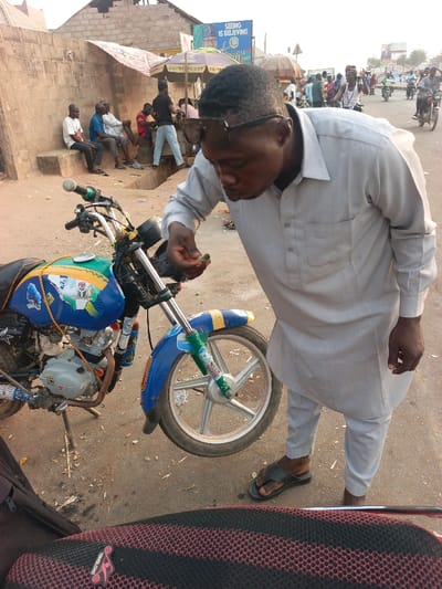 An outdoor scene in Alizaga, Nigeria, depicts a man in a light-colored traditional tunic and trousers, with sunglasses perched on his head, leaning over a blue and yellow decorated motorcycle parked on its kickstand. He is observing a small, dark object held in his left hand. His right hand is at his side, and he wears dark sandals. The motorcycle features a five-spoke silver front wheel and various decals, including a visible Nigerian flag motif on its tank. The immediate ground is unpaved and dusty, transitioning to a paved road further into the background. To the left, several men are seated on a low concrete wall. The background reveals a bustling street environment with multiple additional motorcycles, pedestrians, and market stalls under umbrellas, one displaying "SEEING IS BELIEVING GOMBE 2027." Overhead electrical lines and a utility pole are also visible. The scene is illuminated by daylight.