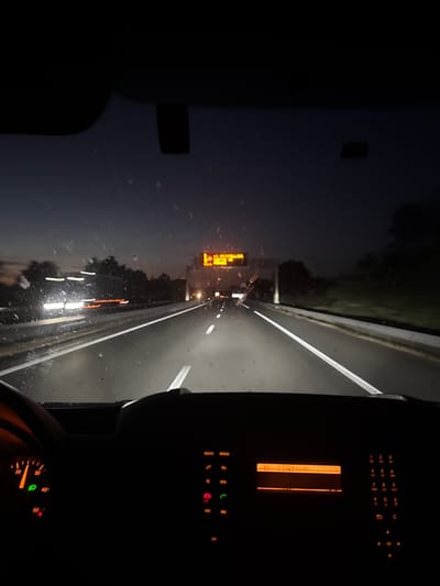 The image is taken from inside a vehicle at dusk, looking forward through the windshield onto a highway. The road has multiple lanes marked with white lines, and oncoming headlights are visible as streaks of light, indicating movement. Trees and foliage line the sides of the highway, and a dark sky suggests it is either sunrise or sunset. An overhead digital sign displays yellow text, possibly indicating traffic information or a destination. The dashboard of the vehicle is visible in the foreground, with a speedometer showing a reading around 30 and a radio display showing "10:23:01:45". The windshield is dirty, with streaks and smudges visible. There is no indication of the specific location within the city of Fontcouverte, France.