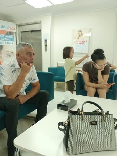 This media file captures a candid scene in what appears to be a waiting area, likely a clinic or doctor's office in Ankara, Turkey. The setting is modern and clean, with turquoise seating and bright, artificial lighting from above, suggesting it's daytime.

The primary subjects are three people. On the left, an older man with graying hair is seated, leaning forward with his chin resting on his hand, seemingly lost in thought or observation. He is wearing a white short-sleeved shirt with a pattern of blue fish. To his right, a young child with dark hair, some of which is dyed pink, is standing and reaching towards another person. This second person, a woman with her hair tied up in a bun, is seated and looking down at her phone, her hand supporting her chin. She is wearing a dark brown t-shirt and glasses. Another person, partially visible behind her, is also seated.

In the foreground, a white table holds a box of tissues and a stylish, black and white patterned handbag with gold hardware. The overall mood is one of quiet waiting, with the child's active gesture contrasting with the stillness of the adults.

Visible text includes "vidi göz" which translates to "vidi eye" and appears on advertisements related to eye care. Another poster, also in Turkish, advertises a solution for "KURU GÖZÜNÜZ OLABİLİR!" meaning "Your eyes may be dry!". Other text on the posters relates to eye treatments like "TOUCH LASER" and mentions "YAKIN VE ASTIGMAT TEDAVİSİNDE," which means "In the treatment of myopia and astigmatism." The brand name "Papia" is also visible on the tissue box.