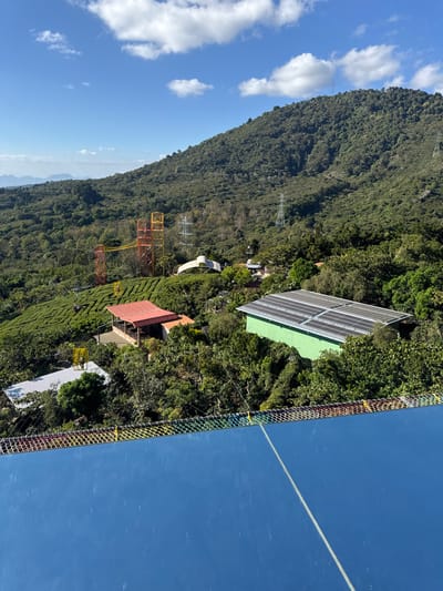 A landscape with a lush green mountain under a partly cloudy sky. Solar panels and several buildings are nestled within the trees. An orange metal structure and power lines are visible on the mountainside. The foreground features blue, potentially reflective material with netting.
