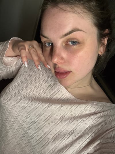 A close-up portrait features a young person, likely female, from the chest up, positioned at an angle with her head tilted. Her striking blue eyes gaze directly at the camera. She has fair skin, dark hair, and subtly defined eyebrows, with a nose piercing visible. Her lips are slightly parted, and her expression is neutral yet engaging.

She wears a light-colored, possibly light pink or beige, long-sleeved top characterized by a detailed, textured weave. The fabric displays alternating vertical bands of fine stripes and small diamond-shaped eyelet patterns. A delicate silver chain necklace is visible around her neck.

Her left hand is gently placed near her face, with manicured nails, predominantly in a light, solid color, while one nail features an intricate white design. A ring with a clear stone is worn on her left ring finger.

The background is dark and out of focus, indicating an indoor setting. The lighting is soft and appears to be ambient or artificial. The image gives the impression of a self-portrait. No text is visible. The location context provided is Chornomorskoe, Ukraine.
