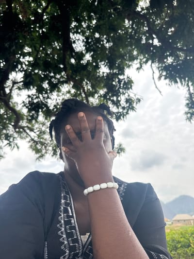 The media file shows a person with dark skin, seen from a low angle outdoors. The person is wearing a black garment with white embroidered patterns and a white beaded bracelet on their wrist. Their face is partially obscured by their hand, which covers their eyes. Their hair is styled in braids. The background features the dappled light filtering through green foliage of trees, suggesting an outdoor setting with abundant vegetation. The sky above is overcast with grey clouds, indicating a cloudy day. In the upper right portion of the image, hints of buildings can be seen, suggesting a semi-urban or village environment. The overall impression is one of a candid, possibly reflective moment captured outdoors in Jawu, Nigeria, under a cloudy sky.