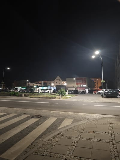 The image shows a street scene at night in Stargard, Poland. A road with a pedestrian crossing is in the foreground. Across the street, there are several buildings, including a hotel with a sign that says "HOTEL" in large letters. Other buildings house shops and businesses, one with a large sign that reads "CHIŃSKIE CENTRUM HANDLOWE" (Chinese Shopping Center). Several cars are parked in front of the buildings and on the street. Streetlights illuminate the scene. The sky is dark.