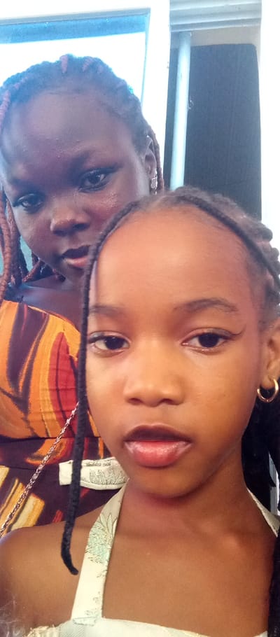 The scene, located in the city of Udi, Nigeria, features a close-up, vertical portrait of two individuals. In the foreground, a young female child with dark skin and dark braided hair looks directly at the camera. She wears a light-colored, possibly halter-neck, top with a subtle pattern, a small gold hoop earring in her right ear, and dark eyeliner. Her lips are slightly parted. Behind her and to the left, an older female, likely a teenager or young adult, with dark skin and reddish-brown braided hair is partially visible. She wears a top with an orange, brown, and black abstract pattern and a silver-colored earring in her left ear, looking slightly to the viewer's left. A thin silver chain is visible near the foreground subject's left shoulder. The background is indistinct, showing a light-colored vertical structure and dark areas, consistent with an indoor environment, with a bright area resembling a window or light source at the top.