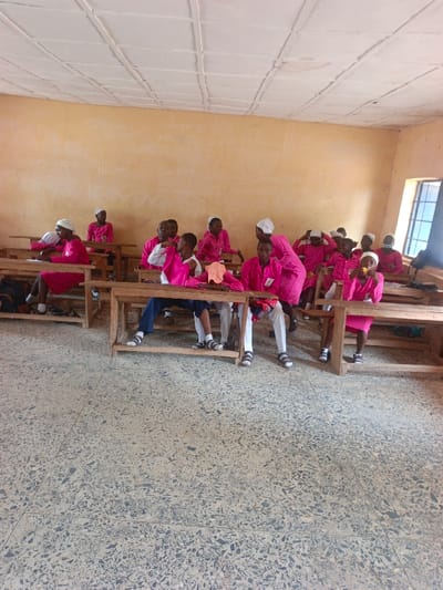 The image displays an interior room, likely a classroom or educational setting, located within a facility in Mada, Nigeria. Approximately 18 individuals, appearing to be adolescents and young adults, are present. Most individuals wear matching bright pink long-sleeved uniforms and white head coverings. Some also wear white trousers, while one individual wears blue trousers. They are seated at simple wooden desks and benches arranged in rows. In the left foreground, a person in a pink uniform is seated, engaged with items on their desk. In the center, two individuals sit at a desk with an orange fabric item resting on it; one wears a white shirt and blue trousers, the other a pink uniform and white trousers. A person in a pink uniform stands and leans over a desk in the mid-right. To the right, another person in a pink uniform consumes a yellow item. Several individuals have identification badges pinned to their uniforms. The walls are painted a light tan color with faint white markings. The ceiling is white with a visible grid pattern. The floor is light grey terrazzo with dark aggregate. A dark-framed window on the right wall admits natural light and offers a view of a bright exterior.