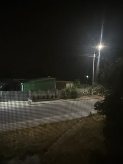 The image depicts a nighttime scene viewed through a screen, likely from a window, showing an outdoor area adjacent to a street. In the background, several large, rectangular structures, possibly shipping containers, are visible, one of which is dark green. A street light illuminates a portion of the scene with a bright, vertical beam. Trees and foliage are present, especially on the right side of the image, obscuring some of the background. A road is visible in the middle ground, with a car parked or driving on it. The foreground shows a grassy or dirt area and a textured surface, possibly pavement or a sidewalk, also seen through the screen mesh. The overall environment appears to be an industrial or semi-urban area.