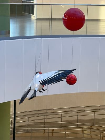 This image features a large, geometric sculpture of a bird in flight, likely suspended indoors or in a sheltered outdoor area. The bird sculpture is predominantly white with sharp, angular wings and tail feathers in black and grey. It has a red beak and a red, spherical object is perched on its wing, appearing to be a stylized ornament or decorative element.

To the left of the bird sculpture, a large, red, spherical object is visible, also possibly suspended. It reflects the surrounding environment, showing some light sources and building elements.

The background is a mix of modern architecture. On the left, there's a large pane of reflective glass, showing muted reflections of a building's interior or exterior, including what appear to be window frames. To the right of the bird sculpture, there's a smooth, white, paneled wall with horizontal lines. Further to the right, a section of a wall covered in small, light-colored tiles is visible, with a structure of vertical and horizontal metal bars.

The lighting suggests it's daytime, with bright illumination that creates reflections and highlights on the shiny surfaces of the sculptures and the glass. There are no people visible in the image. The overall impression is of a public art installation within a contemporary architectural setting. The location context provided is Borisova Griva, Russia.
