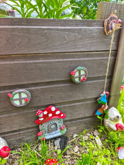 This is a close-up, eye-level, outdoor photograph of a wooden planter box decorated with gnome and mushroom figurines. The planter box is made of dark brown, horizontal wooden planks. Attached to the side of the planter are several whimsical decorations.
At the top right, a gnome figurine with a brown hat and blue tunic is attached to a wooden post, appearing to hang by a twine. Below this, three smaller gnome figurines are hanging from the same twine, one in a blue shirt, another in a brown shirt, and the third's attire is partially obscured.
On the planter wall itself, there are two round, green-topped window decorations, each adorned with red and white spotted mushrooms. A miniature mushroom-shaped house with a "Welcome" sign above its door is also affixed to the planter.
In the foreground, on the ground next to the planter, are more garden decorations including a white hedgehog figurine holding a red apple and several small red and white spotted mushroom caps scattered amongst the green grass and soil.
The background shows lush green foliage and hints of trees, suggesting a garden setting. The photograph is well-lit, with natural daylight. The location is identified as Puchezh, Russia.