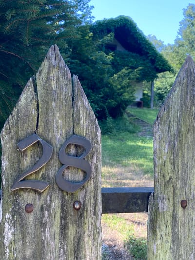 The image shows a weathered wooden gate with the numbers "28" attached to it. The gate is made of two pointed wooden posts, each showing signs of age and moss growth. The numbers are metallic and dark in color. In the background, a house covered in ivy is visible, with a pathway leading up to it. The setting appears to be a rural or suburban area with trees and green grass. The location is the city of Pressbaum, Austria.