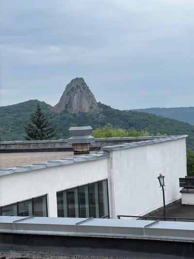 The image shows a view of the Beshtau mountain in Zheleznovodsk, Russia, from the roof of a building. The mountain is a prominent geological formation with a rugged, rocky peak covered in sparse vegetation. It is surrounded by a dense, green forest that covers the rolling hills. In the foreground, the roof of a white, modern building is visible. The roof has a flat surface with a gravel finish in some areas and is bordered by a metallic flashing. A large ventilation unit is situated on the roof, along with a lamppost with an ornate design. The sky is overcast and appears grey, suggesting a cloudy or possibly rainy day. The overall scene depicts a juxtaposition of natural landscape and urban infrastructure.