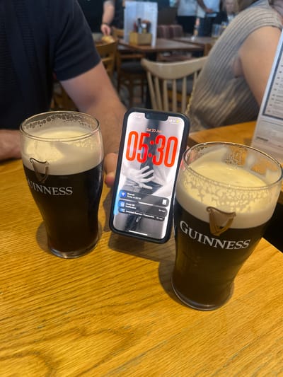 The image depicts a close-up, eye-level shot of two glasses of Guinness on a wooden table. A smartphone is positioned between the glasses, displaying the time 05:30 and notifications. The background is blurred but suggests an indoor setting, possibly a pub or restaurant, with other patrons visible. The lighting appears to be artificial and ambient. No specific landmarks or architectural details of Uttlesford are visible, making it impossible to pinpoint the exact location within the district.