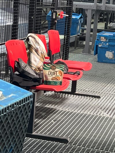 The image shows a seating area with red plastic chairs. On one of the chairs, there is a black bag, a plaid blanket draped over the back, a bottle of red liquid, and a bag of "Flint CRAFT" chips. The seating area appears to be in a large, industrial-like space with metal grating flooring and fencing in the background. There are blue bins visible in the background. The setting is likely a public area or waiting zone within the city of Nerubais'ke, Ukraine.