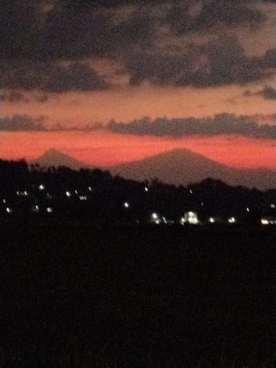 The image depicts a rural landscape at dusk, likely in Menjing, Indonesia. Two prominent, conical mountains are silhouetted against a vibrant, fiery orange and pink sunset. Dark, textured clouds fill the upper portion of the sky, adding depth and drama to the scene. Below the mountains, a dark band of trees or hills runs horizontally across the mid-ground. Scattered lights, appearing as soft, out-of-focus glows, are visible amongst this dark terrain, suggesting a village or scattered settlements. The foreground is a completely dark, unlit expanse, possibly a field or undeveloped land. The overall impression is one of tranquility and the natural beauty of the Indonesian countryside as day transitions into night. The image has a grainy quality, which might be due to low light conditions or the photographic equipment used. No people or specific objects are clearly discernible.