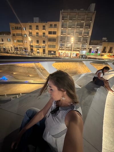 The image depicts two women in an outdoor urban setting at night. One woman is in the foreground, with her head bowed and to the side, wearing a white tank top and blue jeans. She has a starfish-shaped earring. The second woman is in the midground, sitting with her legs crossed and looking to her left. She is wearing a white top and dark shorts.

The background shows a multi-story building with many windows, illuminated by artificial lights, suggesting it's a commercial or residential area. There are also lights from shops or restaurants visible on the building's lower floors. The ground appears to be a paved area, possibly a walkway or plaza, with a decorative element that reflects light, possibly a water feature or textured surface. The overall lighting suggests it is nighttime, and the scene appears calm. The image is oriented vertically, with the top of the frame appearing on the left. No visible text is discernable in the image.