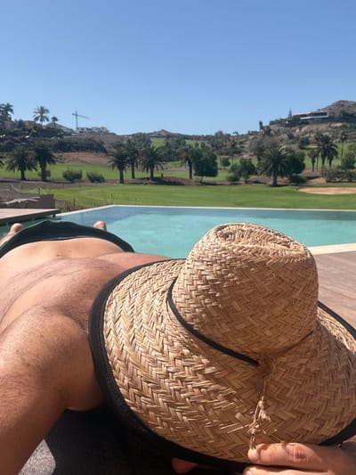 A person is lying on a lounge chair by a swimming pool, wearing a straw hat that covers their face. The person is wearing dark colored swim trunks. The swimming pool is turquoise and overlooks a golf course with palm trees and hills in the background. In the distance, a construction crane is visible. The sky is clear and blue. The image appears to be taken in the Urbanización Salobre Golf, Spain.