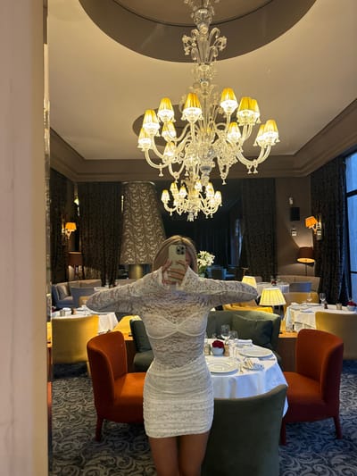 A young woman, wearing a white lace dress and holding a smartphone, is standing in a restaurant in Ouled Jelal, Morocco (31.663, -7.872). She is facing the camera and taking a selfie, with her left hand resting on her hip and her right hand holding the phone. The restaurant is luxuriously furnished, with a large chandelier hanging from the ceiling, a white tablecloth on the table, and several chairs scattered around. The environment is well lit and the walls are painted a light cream color. The woman is standing between two red leather chairs, and a table with a white tablecloth and a glass of water. The table is set with a white napkin and a set of silverware. There are more tables and chairs in the background, suggesting the restaurant is full of patrons.