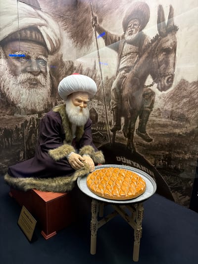 This vertical indoor image displays a museum exhibit featuring a life-size mannequin of Nasreddin Hoca, a renowned figure in Turkish folklore. The mannequin portrays an old man with a long, flowing white beard and a serene, wise expression. He is adorned in traditional attire, including a distinctive white pleated turban with a red cap, a dark purple robe, and a matching fur-trimmed cloak. He is seated on a dark wooden box, appearing to reach towards a large, golden-brown dessert, likely baklava or a similar pastry, arranged in a diamond pattern on a silver platter. The platter rests on a small, intricately carved wooden stand.
The background features a large, sepia-toned mural or artwork, visible through a reflective surface. This artwork depicts scenes related to Nasreddin Hoca, including an illustration of a man (presumably Hoca) riding a donkey, and a detailed close-up portrait of an old man's face. Partially visible text on the mural includes "DÜNYANIN" (of the world).
At the base of the exhibit, a small gold-colored plaque clearly identifies the figure as "NASREDDİN HOCA." Further text on the plaque, though partially obscured, includes references to "Ladik, Eskişehir," grounding the exhibit within the local context of Eskisehir, Turkey. The display effectively combines historical representation with a symbolic culinary element, likely a nod to Turkish culture. The scene is illuminated by indoor artificial lighting, which creates minor reflections on the protective surface covering the background mural.