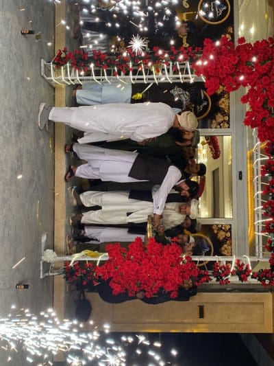 The image shows a group of men standing in front of a building decorated with red flowers and string lights. Some men are wearing traditional clothing, including white shalwar kameez and head coverings. There is a building behind the men with windows and what appears to be decorative elements. Sparklers are going off on the ground in the foreground. The location is possibly قندهار, Afghanistan, based on the presence of men wearing traditional Pashtun clothing.