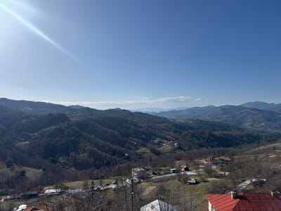 This image captures a panoramic view of Bukova Polyana, Bulgaria, from an elevated vantage point. The scene is dominated by a clear, bright blue sky with a prominent sun flare emanating from the upper left quadrant. The sun's rays extend diagonally across the upper sky.

The landscape consists of undulating hills and mountains that recede into the distance, exhibiting varying shades of green from coniferous forests and bare branches of deciduous trees, indicating a late winter or early spring season. Patches of lighter, possibly agricultural land or cleared areas, are visible on some slopes.

In the midground and foreground, a rural settlement is visible, characterized by scattered individual houses. These structures feature diverse roof types, predominantly red tile and lighter-colored materials. One multi-story concrete structure with exposed framing, possibly under construction, is prominent in the mid-right foreground. Smaller outbuildings and sheds are also present.

A network of unpaved or lightly graveled roads winds through the settlement and ascends into the hills. Utility poles with overhead wires are visible, particularly in the immediate foreground, indicating established infrastructure. No individuals or active human interactions are discernible within the frame. The overall environment appears calm and uninhabited by specific human activity at the time of capture.