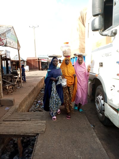 Three women walk along a dirt path next to a large white truck in Gadao, Nigeria. The woman on the left is wearing a dark blue abaya and a patterned scarf, and she is carrying a rolled-up patterned fabric. The middle woman is wearing an orange hijab and a patterned skirt, and she has a round container filled with food balanced on her head. The woman on the right wears a light pink hijab and a matching dress, and she is also carrying a tray of food on her head.

The scene appears to be a market or roadside stall area. To the left of the women are several small structures with corrugated metal roofs, suggesting shops or market stalls. One sign is visible with the text "ADAMU" and "OLAH KALI". In the background, a utility pole stands tall. The ground is dusty and uneven, with a drainage ditch filled with debris running alongside the path. Wooden planks form a makeshift bridge over the ditch. The time of day appears to be daytime, with bright sunlight casting shadows. The overall atmosphere is one of daily activity and commerce.